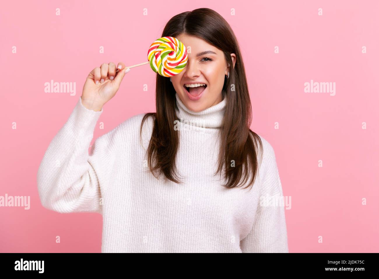 Portrait einer Brünette Frau, die das Auge mit großen leckeren Süßigkeiten bedeckt, die Kamera anschaut, die Zunge herauszeigt und einen weißen Pullover im lässigen Stil trägt. Innenaufnahme des Studios isoliert auf rosa Hintergrund. Stockfoto