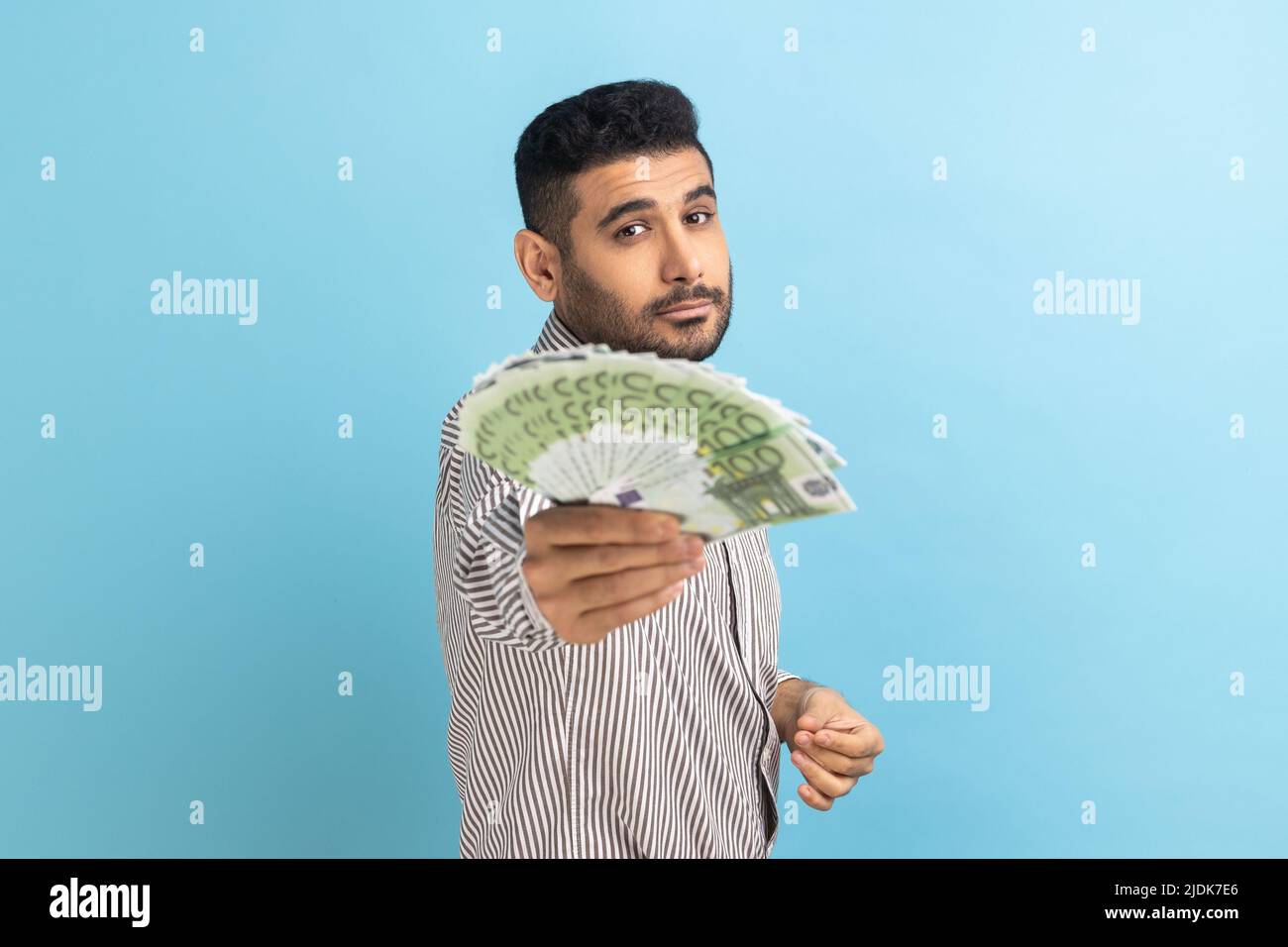 Porträt eines selbstbewussten reichen bärtigen Geschäftsmannes, der Euro-Rechnungen vor die Kamera hält, mit Geld, das im Lotto gewonnen wurde, und einem gestreiften Hemd. Innenaufnahme des Studios isoliert auf blauem Hintergrund. Stockfoto