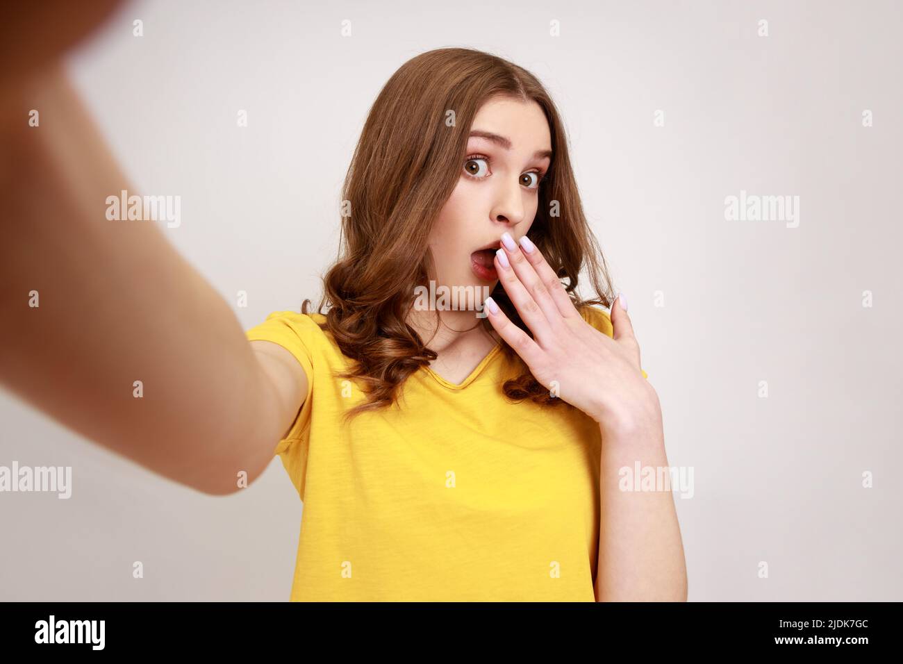 Attraktives Mädchen im Teenageralter, das ein gelbes T-Shirt im Kasaul-Stil trägt, den Mund mit der Handfläche bedeckt, den Blick auf den POV der Kamera und den Blickwinkel des Fotos betrachtet. Innenaufnahme des Studios isoliert auf grauem Hintergrund. Stockfoto