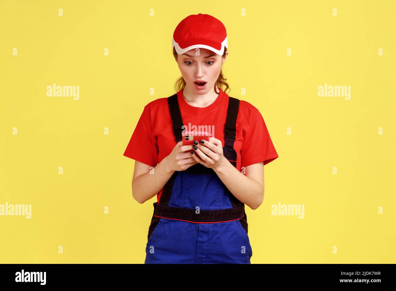 Porträt einer jungen erwachsenen schockierten Arbeiterin, die das Mobiltelefon zur Online-Unterstützung benutzte, positive Emotionen ausdrückte, Overalls und rote Mütze trug. Innenaufnahme des Studios isoliert auf gelbem Hintergrund. Stockfoto