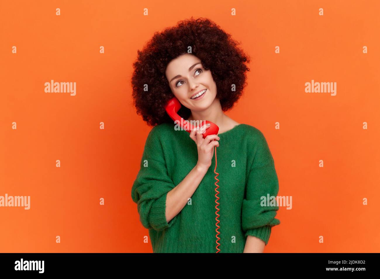 Portrait einer positiven verträumten Frau mit Afro-Frisur in einem grünen Pullover im lässigen Stil, der über ein rotes Festnetztelefon im Retro-Stil und ein Callcenter spricht. Innenaufnahme des Studios isoliert auf orangefarbenem Hintergrund. Stockfoto