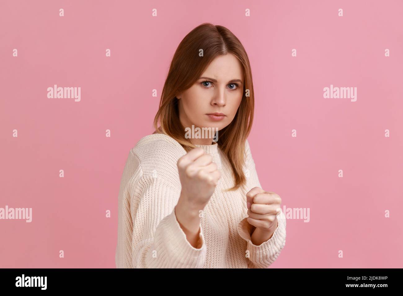 Portrait einer verärgerten selbstbewussten attraktiven blonden Frau, die Fäuste zusammenhält, Boxgesten zeigt und bereit ist, zu schlagen, mit weißem Pullover. Innenaufnahme des Studios isoliert auf rosa Hintergrund. Stockfoto