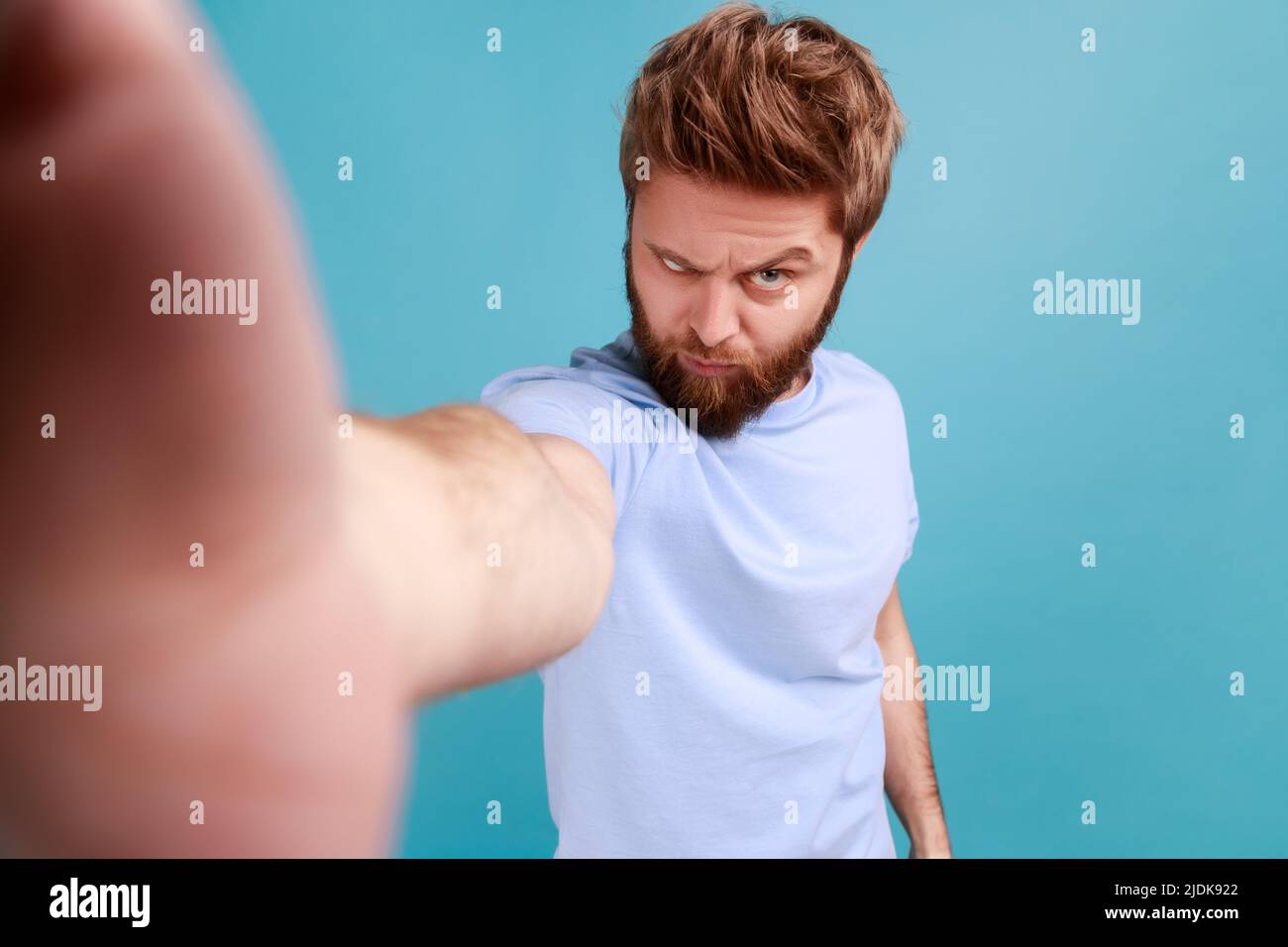 Porträt eines hübschen bärtigen Bloggers, der Selfie macht oder Livestream sendet, die Kamera mit wütendem, aggressivem Ausdruck und einem lustigen Gesicht anschaut. Innenaufnahme des Studios isoliert auf blauem Hintergrund. Stockfoto