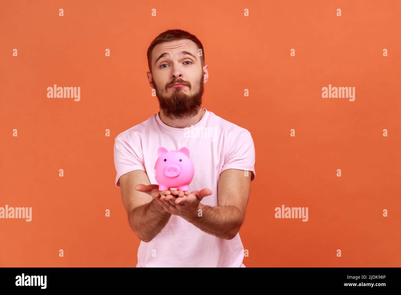 Porträt eines traurig frustrierten bärtigen Mannes, der Sparschwein in den Händen hält, unglücklich ist, keine Ersparnisse hat, Trauer ausdrückt und ein rosa T-Shirt trägt. Innenaufnahme des Studios isoliert auf orangefarbenem Hintergrund. Stockfoto
