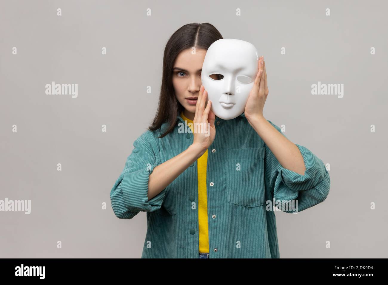 Porträt einer streng herrisch Frau, die die Hälfte des Gesichts mit einer weißen Maske bedeckt, mehrere Persönlichkeitsstörungen und eine Jacke im lässigen Stil trägt. Innenaufnahme des Studios isoliert auf grauem Hintergrund. Stockfoto