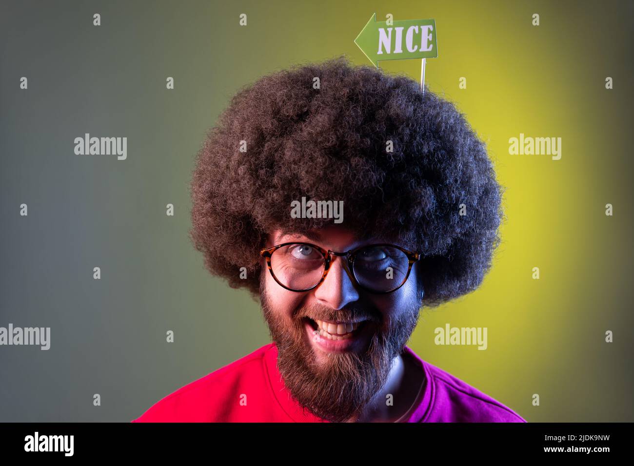 Porträt eines verrückten, glücklichen Hipster-Mannes mit Afro-Frisur, der seinen Urlaub feiert, festliche Stimmung hat und auf Party-Requisiten in seinen Haaren aufschaut. Innenaufnahmen im Studio, isoliert auf farbigem Neonlicht-Hintergrund. Stockfoto