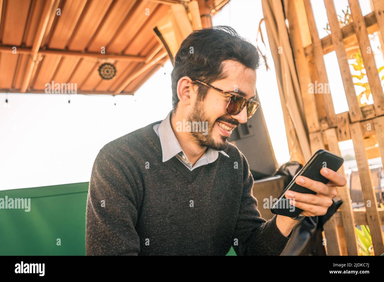 Junger bärtiger Hipster lächelt und schaut in einer Bar auf sein Handy Stockfoto