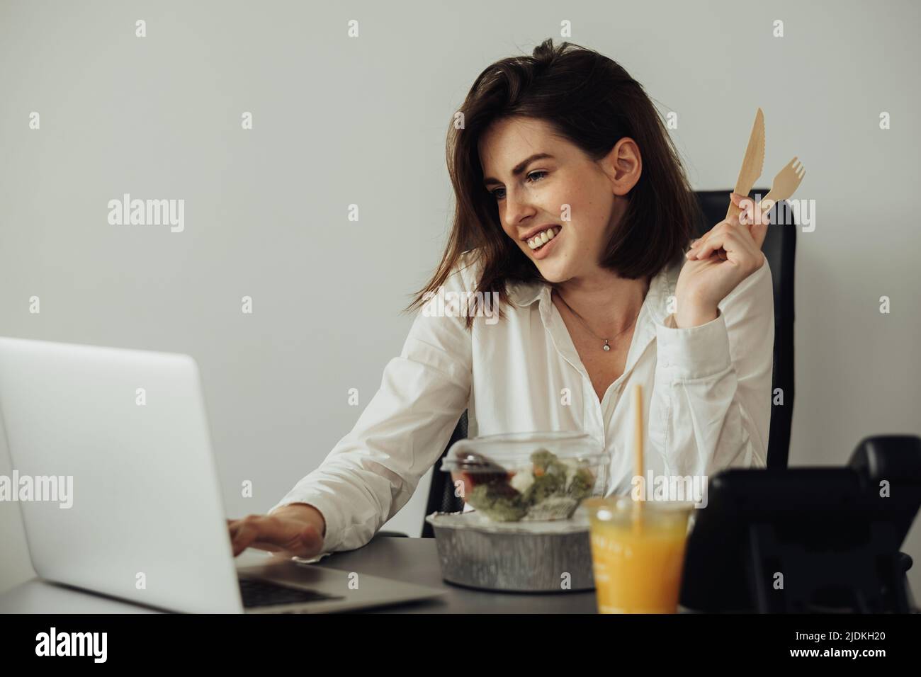 Fröhliche weibliche Angestellte beim Mittagessen im Büro, junge Brünette Frau, die sich auf das Essen am Arbeitsplatz vorbereitet Stockfoto