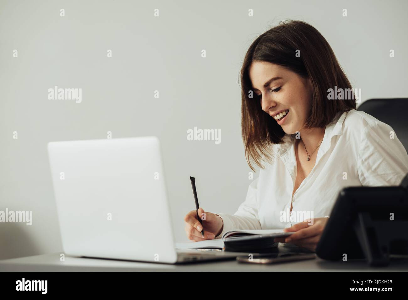 Fröhliche junge Frau, die im Büro arbeitet von Laptop, Buchhalterin, die am Arbeitsplatz ein Notizbuch schreibt Stockfoto