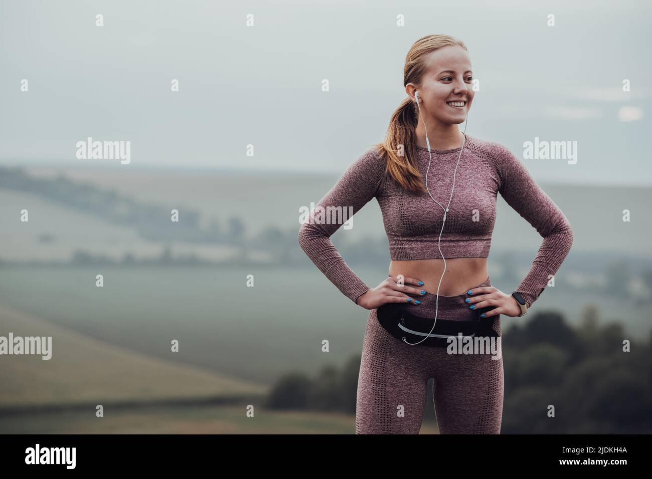 Fröhliche junge kaukasische Frau hält Hände auf Taille und Musik im Freien auf dem Hügel Stockfoto