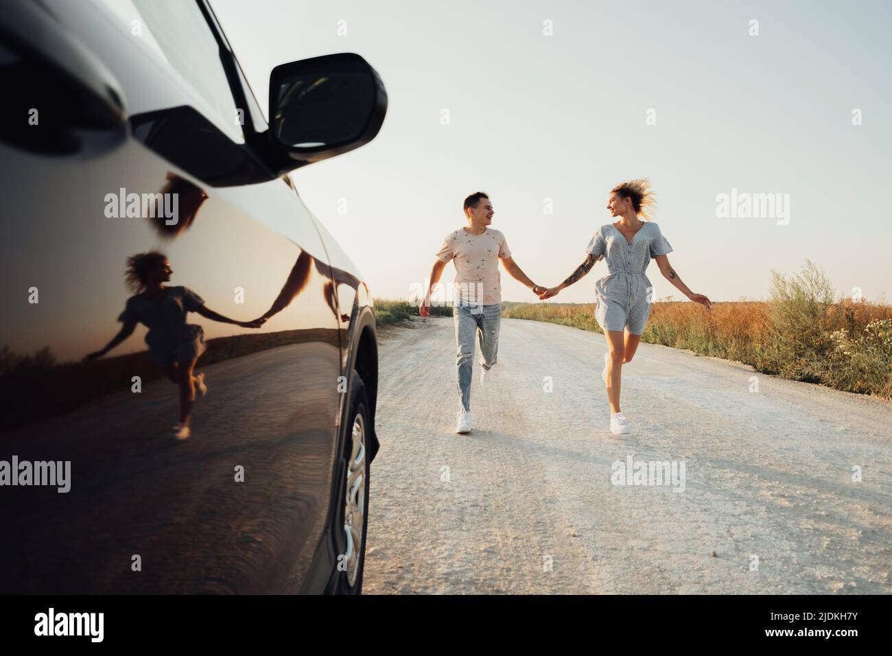 Glücklicher Mann und fröhliche Frau halten sich an den Händen, die in der Nähe des Autos laufen, Junges Paar genießt die Road Trip bei Sonnenuntergang Stockfoto