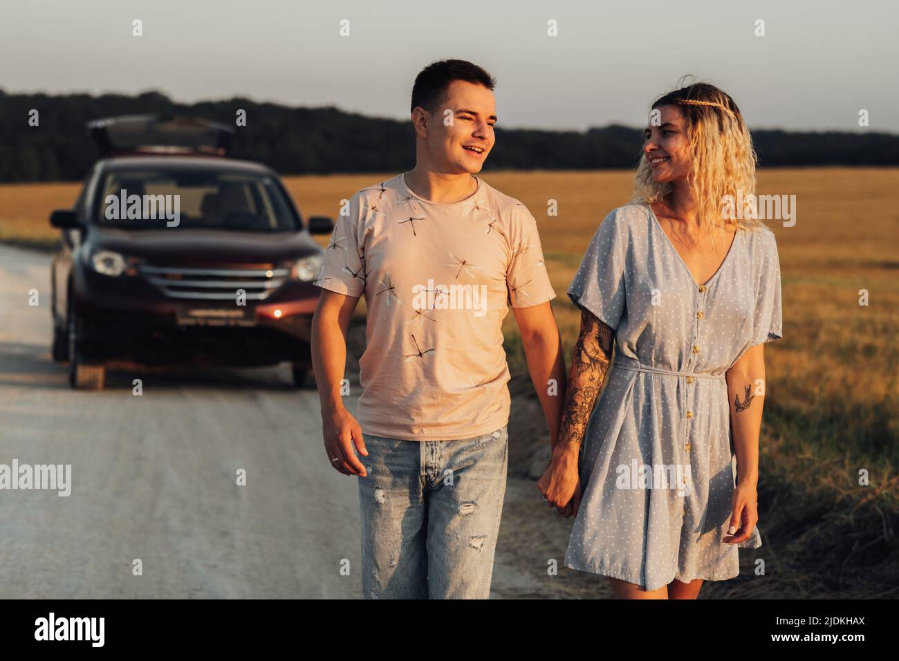 Kaukasischer Mann und Frau halten sich mit den Händen auf den Hintergrund ihres SUV-Autos, Junges Paar genießt Road Trip bei Sonnenuntergang Stockfoto