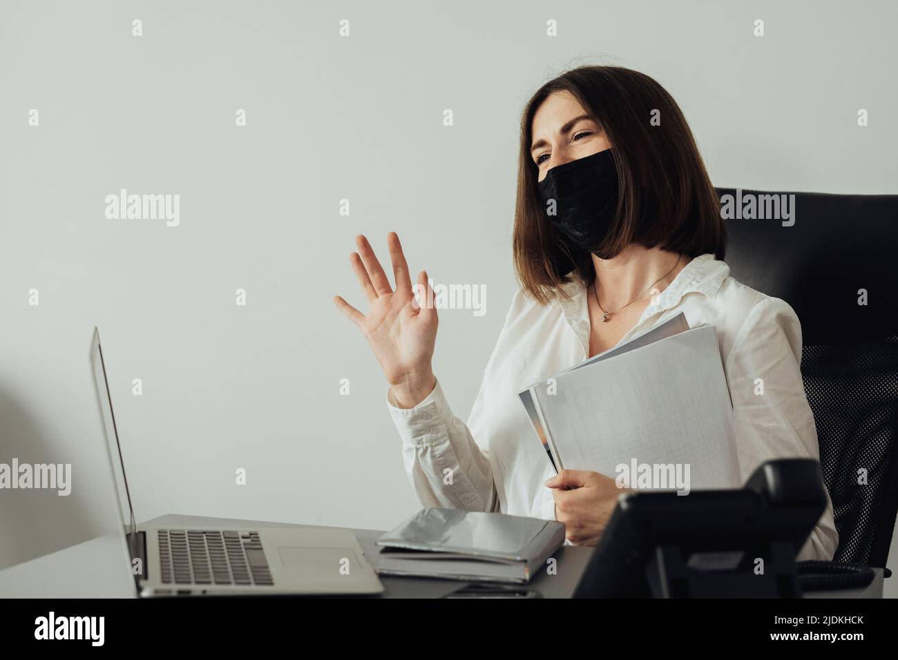 Fröhliche junge Frau in Schutzmaske winkende Hand und grüßt den Klienten, Managerin bei der Arbeit im Büro Stockfoto