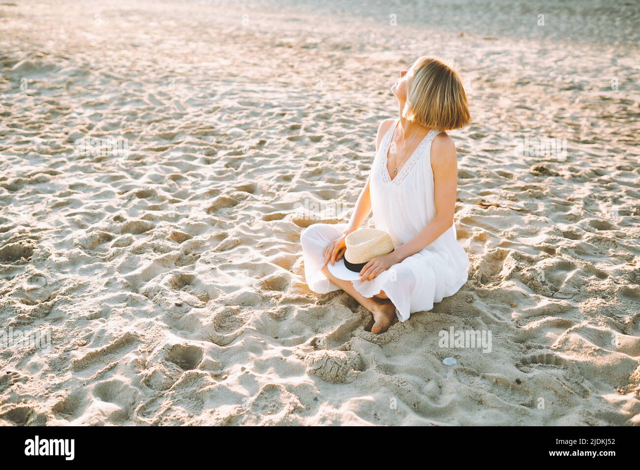 Frau mittleren Alters, die an sonnigen Tagen mit gekreuzten Beinen am Sandstrand sitzt, Hände auf Strohhut legt und die Sonne anschaut. Stockfoto