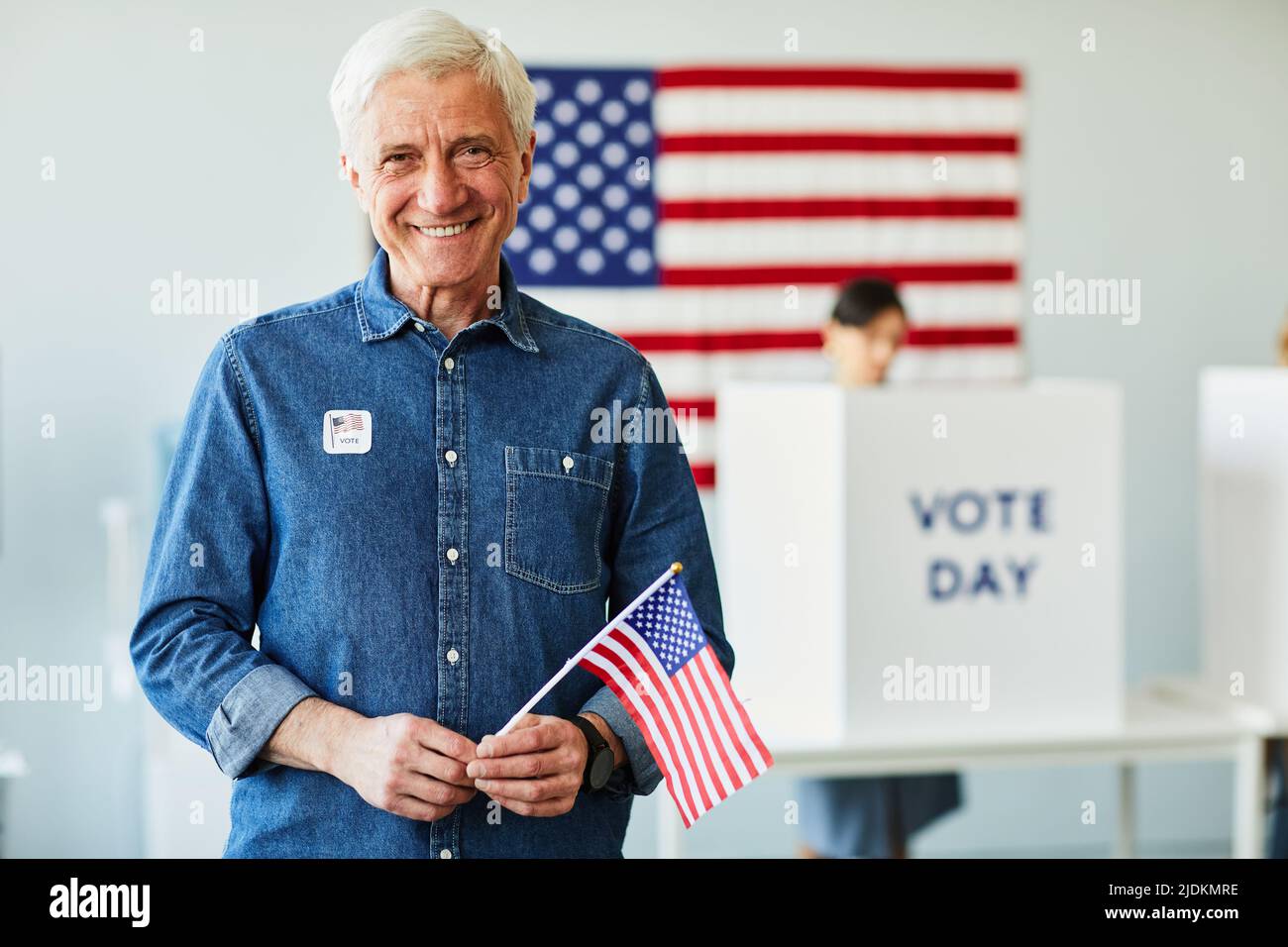 Porträt eines lächelnden älteren Mannes, der am Wahltag die amerikanische Flagge in der Wahlstation hält, in der Taille nach oben, Platz kopieren Stockfoto