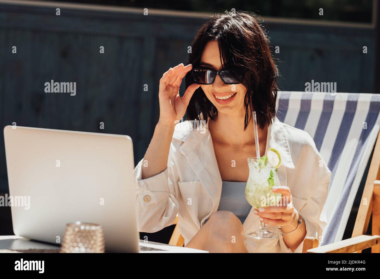 Fröhliche junge Frau in Sonnenbrillen, die Cocktail trinkt und einen Laptop für Videoanrufe verwendet, während sie auf der Terrasse sitzt Stockfoto