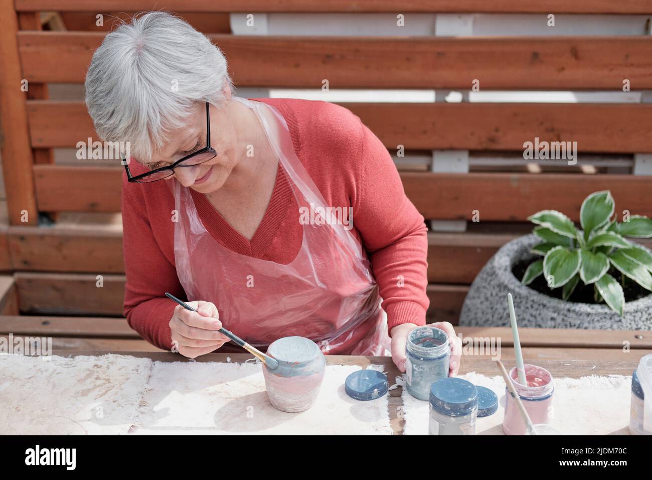 Ältere grauhaarige Frau mit Gläsern, die handgemachte Tonvase mit Farbfarben mit Pinsel malt. Stockfoto
