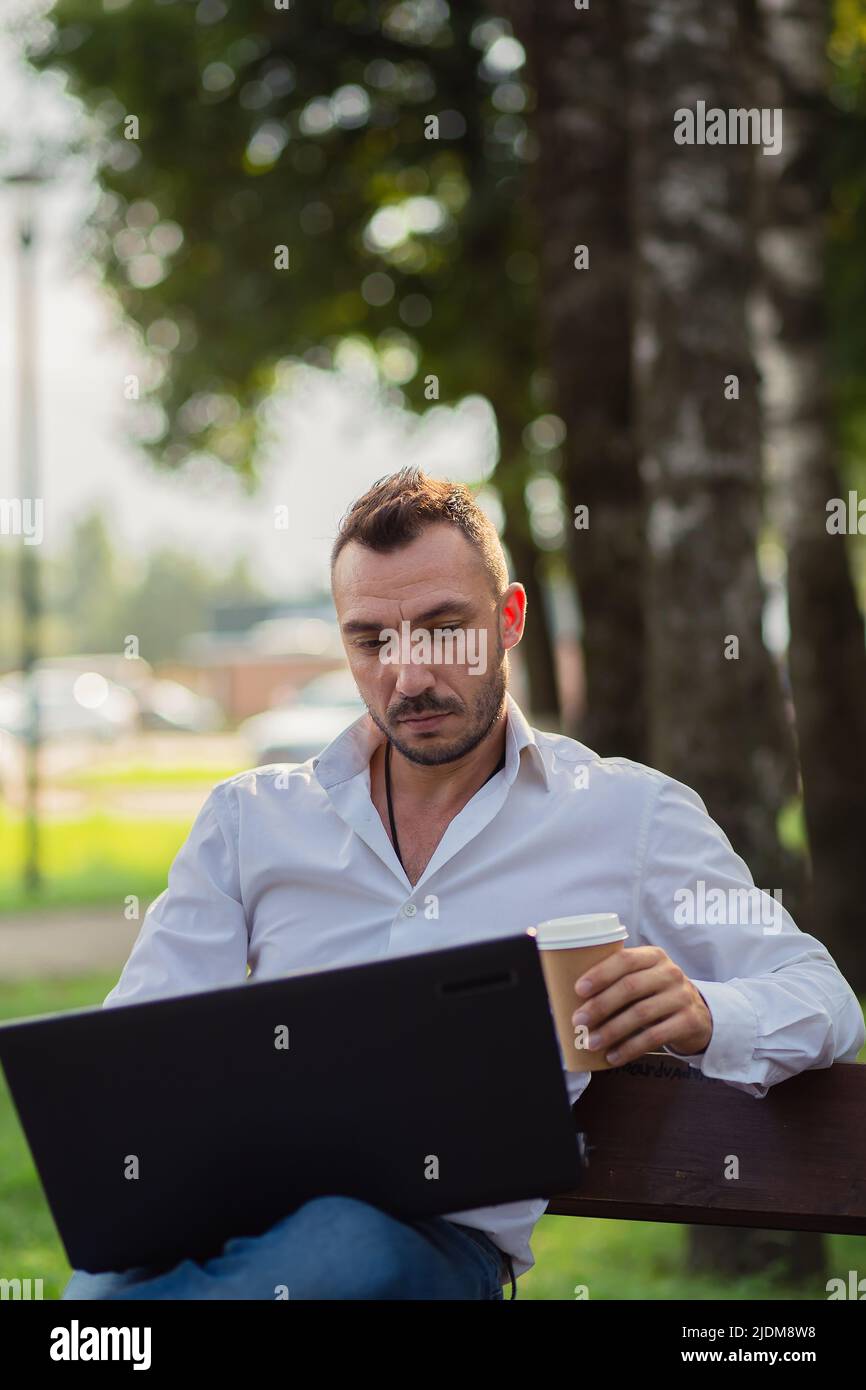 Konzentrierter Mann arbeitet im Park mit einem Laptop, trinkt Kaffee. Ein junger Mann auf einem Hintergrund von grünen Bäumen, ein heißer sonniger Sommertag. Warmes, weiches Licht, Nahaufnahme. Stockfoto