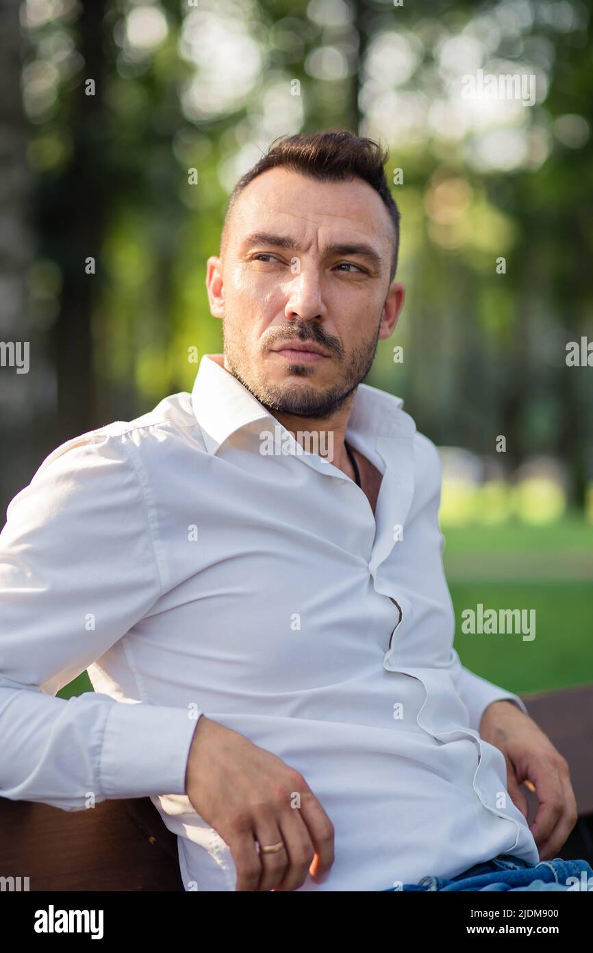 Ein seriöser Geschäftsmann in einem weißen Hemd ruht sich aus, während er auf einer Bank sitzt. Junger Mann im Stadtpark, warmer sonniger Sommertag. Warmes, weiches Licht, Nahaufnahme. Stockfoto Ein seriöser Geschäftsmann in einem weißen Hemd ruht sich aus, während er auf einer Bank sitzt. Junger Mann im Stadtpark, warmer sonniger Sommertag. Warmes, weiches Licht, Nahaufnahme. Stockfoto
