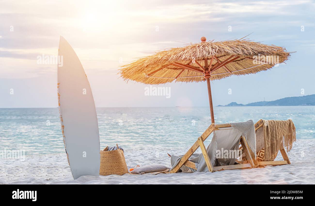 Schöner ruhiger weißer Sandstrand mit zwei Liegestühlen und Strohschirm mit Surfbrett. Romantischer Sommerurlaub auf einer tropischen Privatinsel Stockfoto