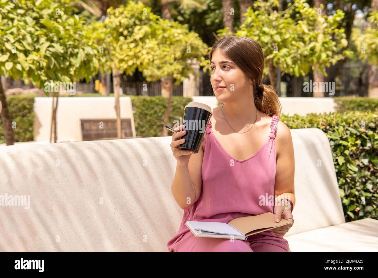 Junge Frau in rosa Sommerkleid, die auf einer Parkbank sitzt, während sie ihren Kaffee trinkt und ihre Notizen in ein Notizbuch schreibt. Stockfoto