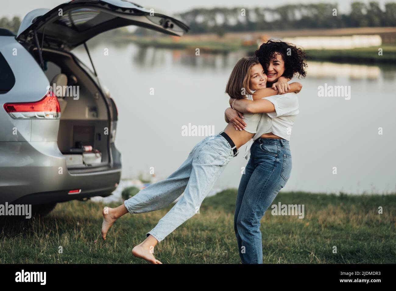 Alles gute zum Muttertag, Frau umarmt sich mit fröhlicher Teenage-Tochter im Freien auf dem Hintergrund von Auto und See Stockfoto