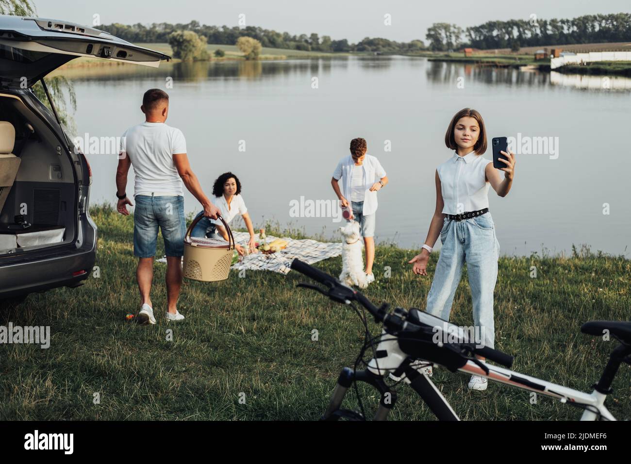 Glückliche vier Mitglieder Familie genießen Wochenende Road Trip mit Minivan Auto, Teenage Tochter Selfie machen, während ihr Bruder spielt mit Haustier Hund am See Stockfoto