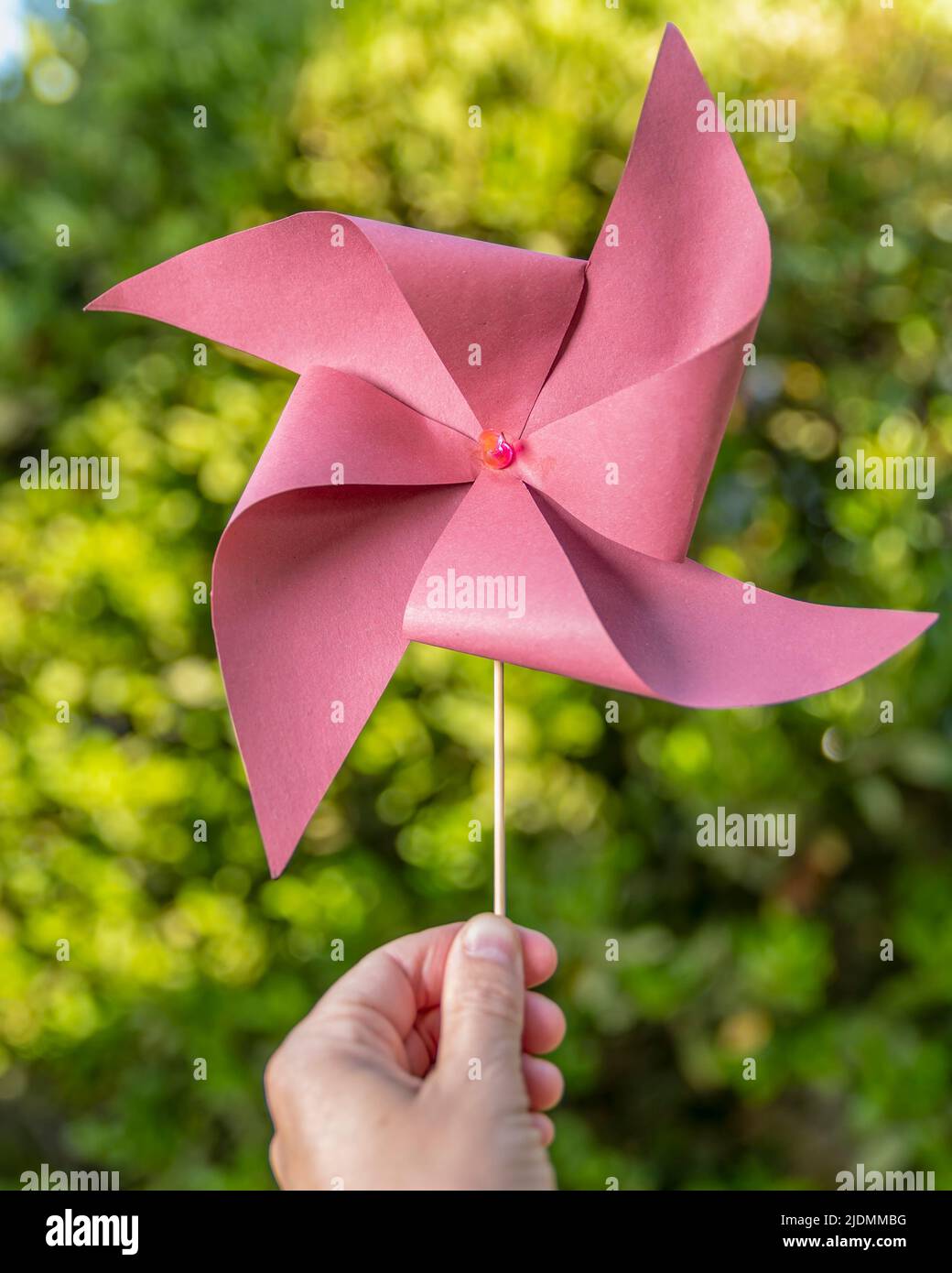 Eine männliche Hand hält ein selbstgemachtes rotes Windrad vor einem verschwommenen grünen Hintergrund mit Bokeh-Lichtern Stockfoto