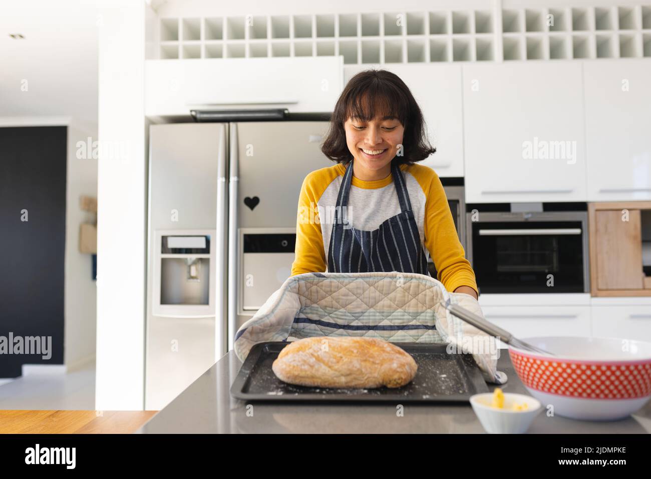 Lächelnde asiatische junge Frau mit kurzen Haaren hält frisch gebackenes Brot auf Kücheninsel Stockfoto