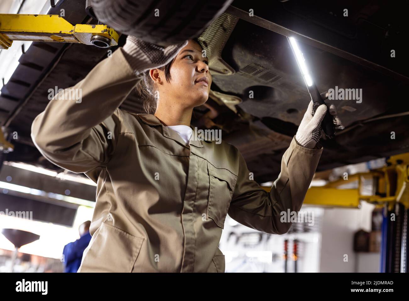 Mittlere Erwachsene asiatische Technikerin, die Beleuchtungsausrüstung verwendet, während sie ein fehlerhaftes Auto in der Werkstatt analysiert Stockfoto
