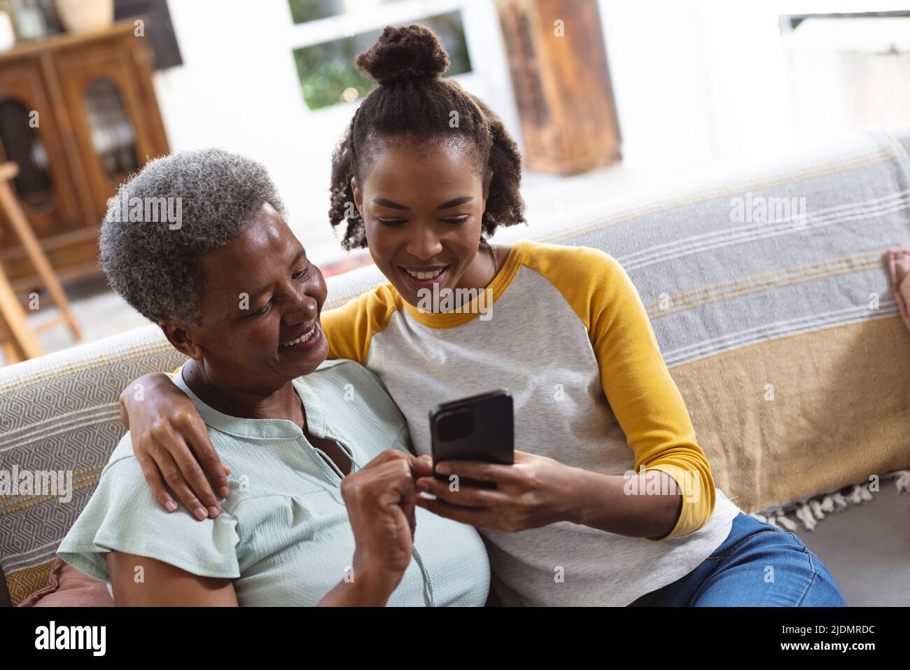 afroamerikanische junge Tochter mit der Hand auf der Schulter der Mutter und Smartphone, während sie auf dem Sofa sitzt Stockfoto