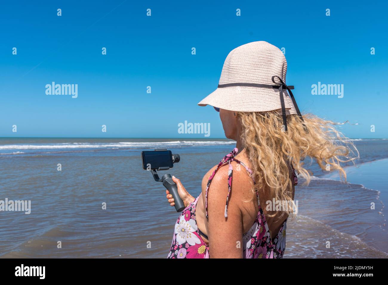 Frau, die ein Mobiltelefon mit einem Stativ benutzt, während der Wind ihre Haare am Strand bewegt Stockfoto