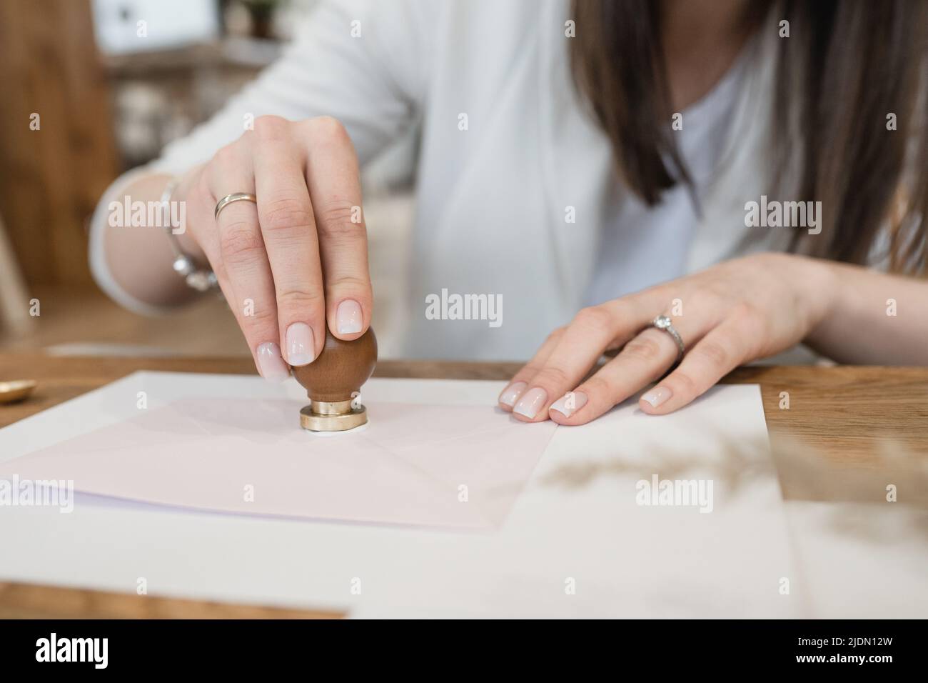 Gepflegte Hände mit Ringen einer nicht erkennbaren Frau, die Wachssiegel mit Stempel auf weißem Umschlag auf Holzschreibtisch macht. Stockfoto