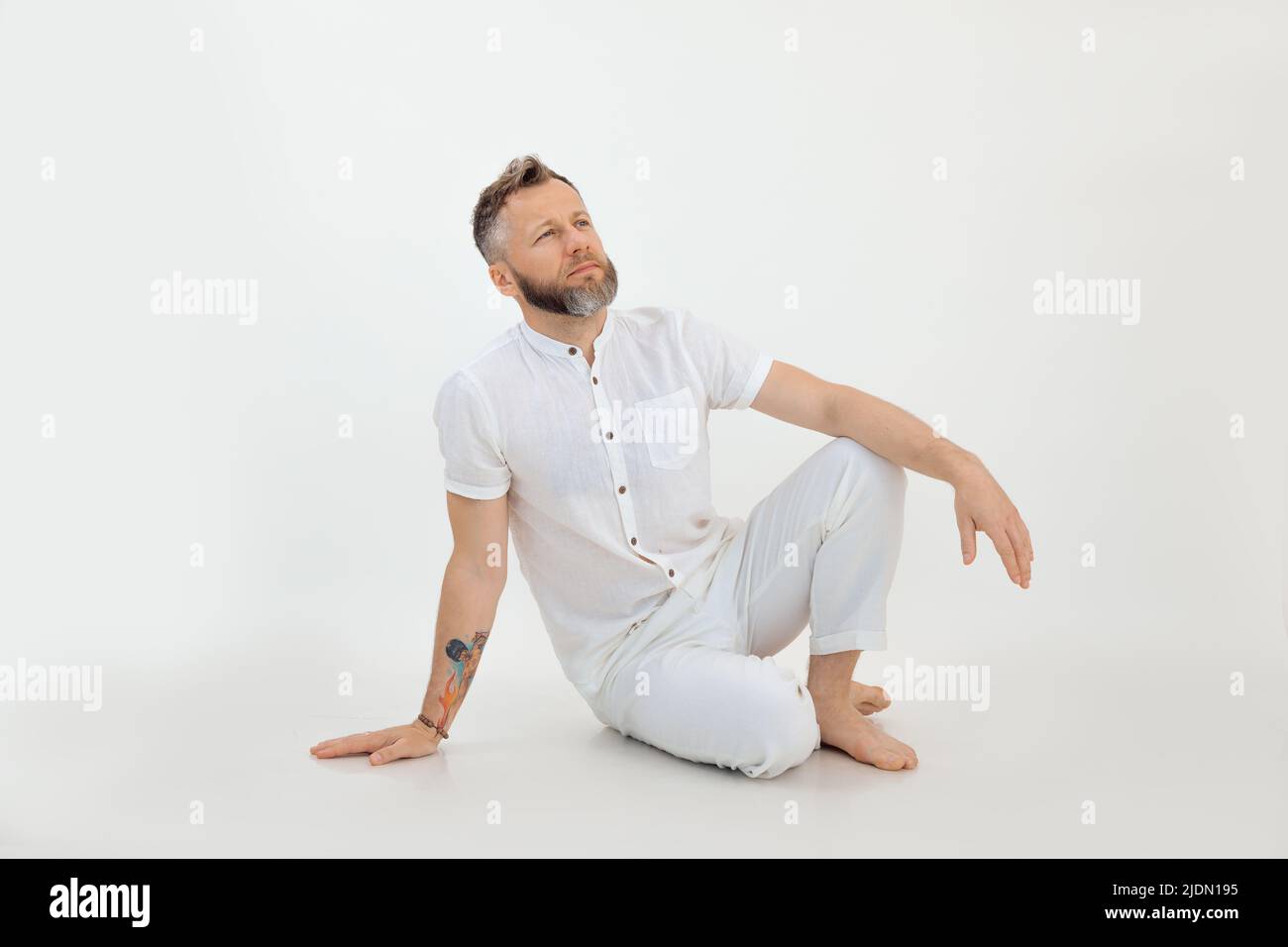 Barfuß nachdenklicher Mann mit Tätowierung, der auf dem Boden in weißem Outfit auf weißem Hintergrund sitzt. Geschäftsgedanken Stockfoto