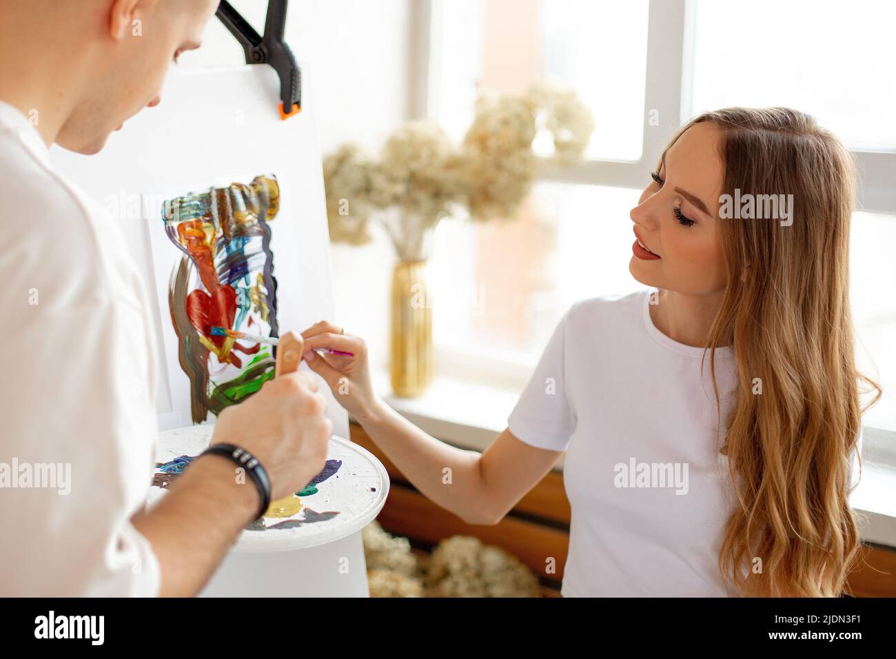 Junges Paar, das sich zusammenzieht. Frau und Mann küssen und halten Pinsel und Pallette mit Farben. Stockfoto