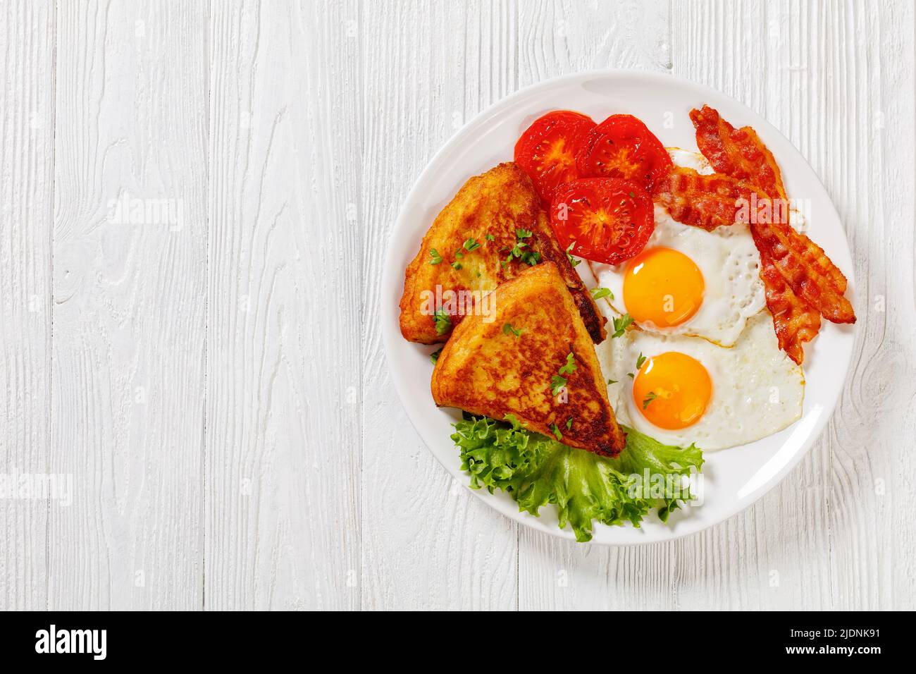 irisches Frühstück mit Kartoffelpüree, Spiegeleiern, Speckscheiben, gegrillten Tomaten und frischem Salat auf weißem Teller auf weißem Holztisch, horizontal Stockfoto