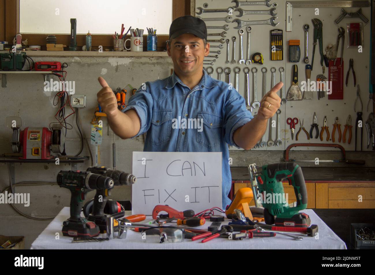 Handwerker mit Daumen nach oben in seiner Werkstatt mit Arbeitswerkzeugen vorne und „Ich kann es reparieren“-Schild im Zusammenhang mit DIY-Arbeit Stockfoto