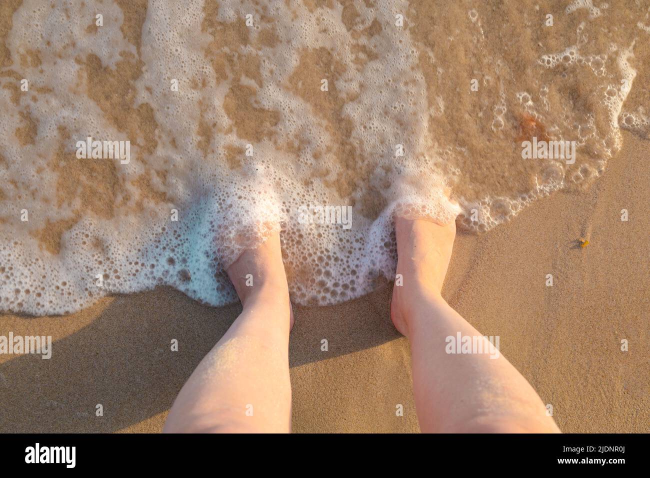 Weibliche Füße im Sand am Meer, bedeckt mit Meereswellen-Schaum. Urlaubsstimmung. Hintergrund Stockfoto