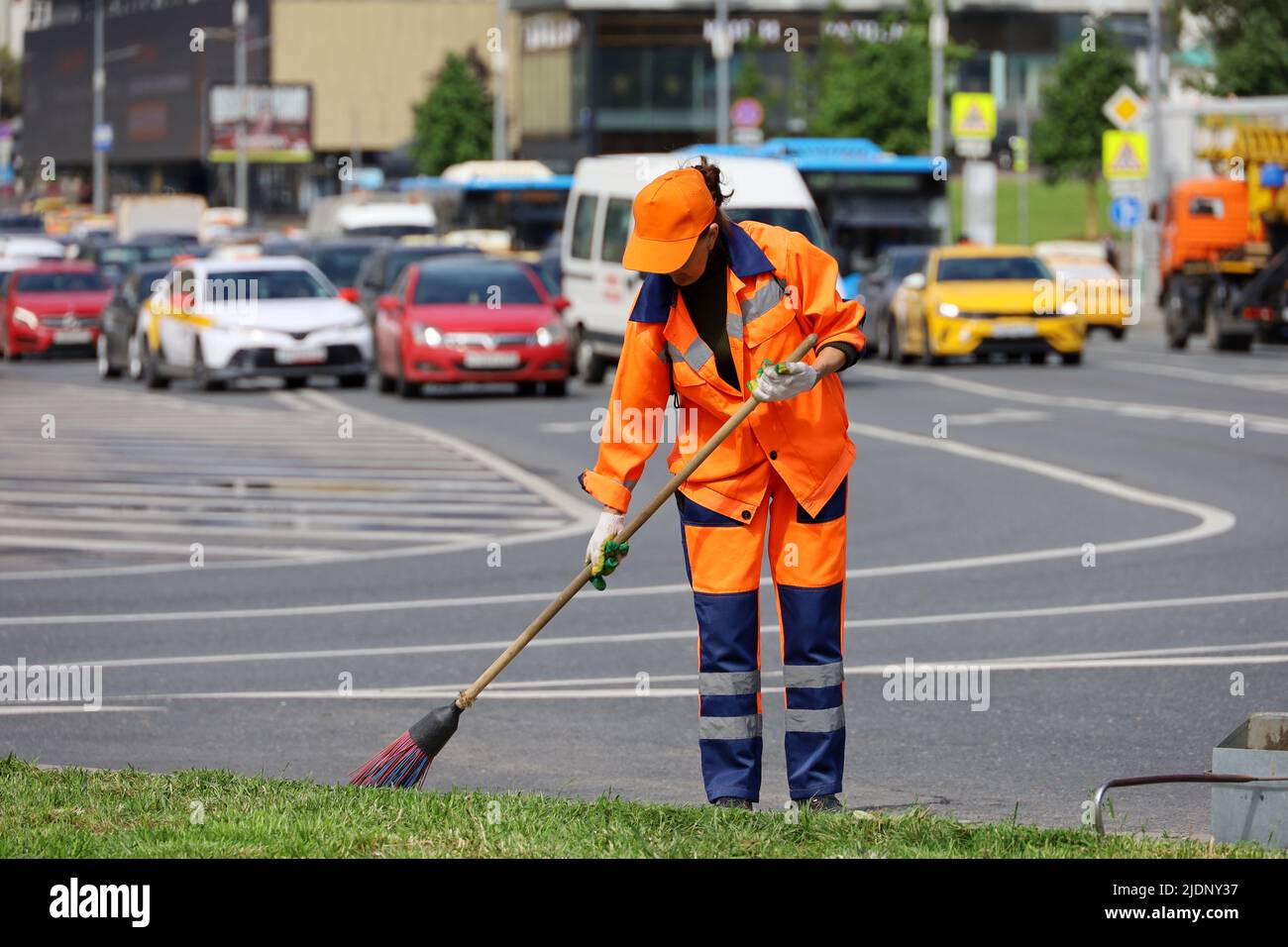 Eine Arbeiterin in orangefarbener Uniform mit einem Besen fegt auf dem Hintergrund eines Autos über die Straße. Straßenreinigung in der Sommerstadt Stockfoto
