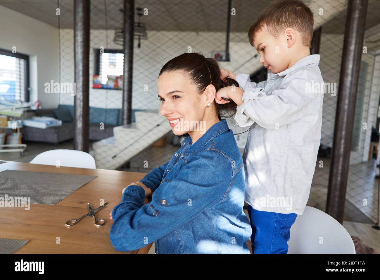 Mutter sitzt am Tisch im Wohnzimmer und lässt ihren Sohn die Haare kämmen Stockfoto