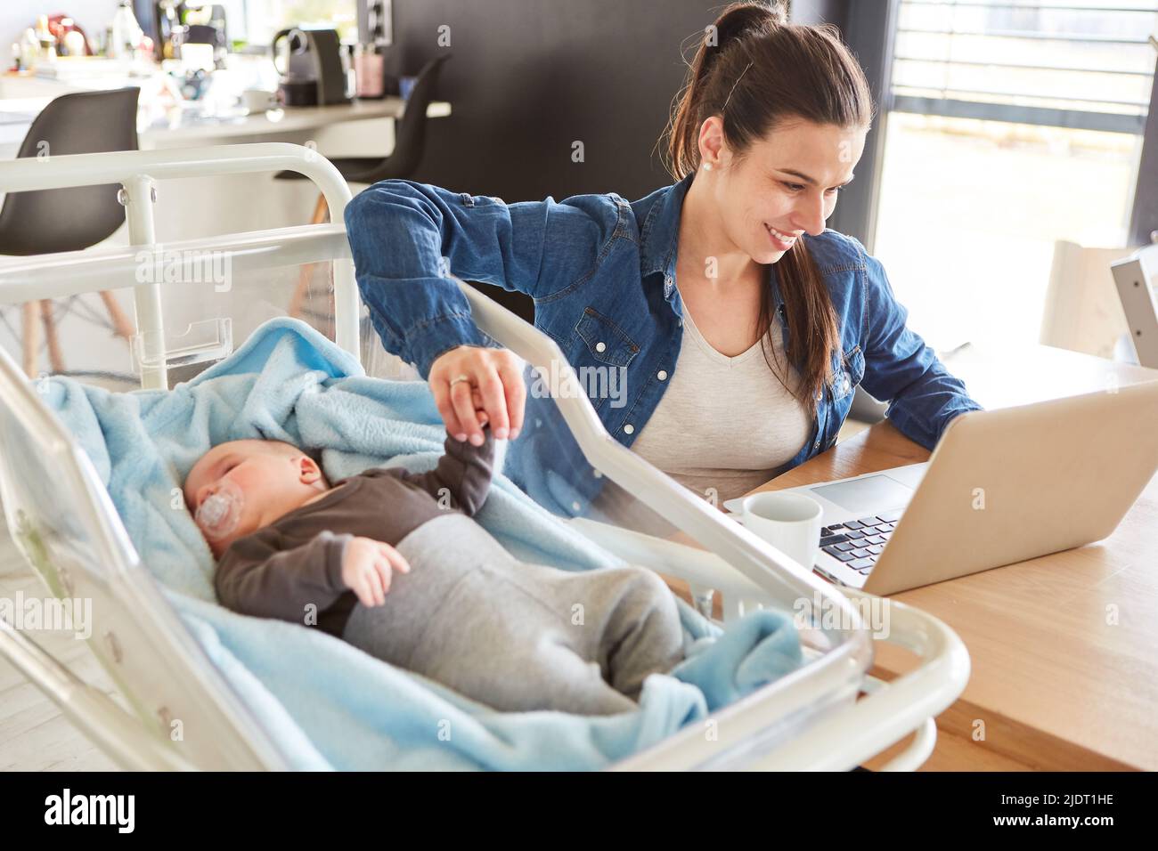 Lächelnde Mutter, die am pc arbeitet und die Hand ihres Babys im Heimbüro neben sich hält Stockfoto