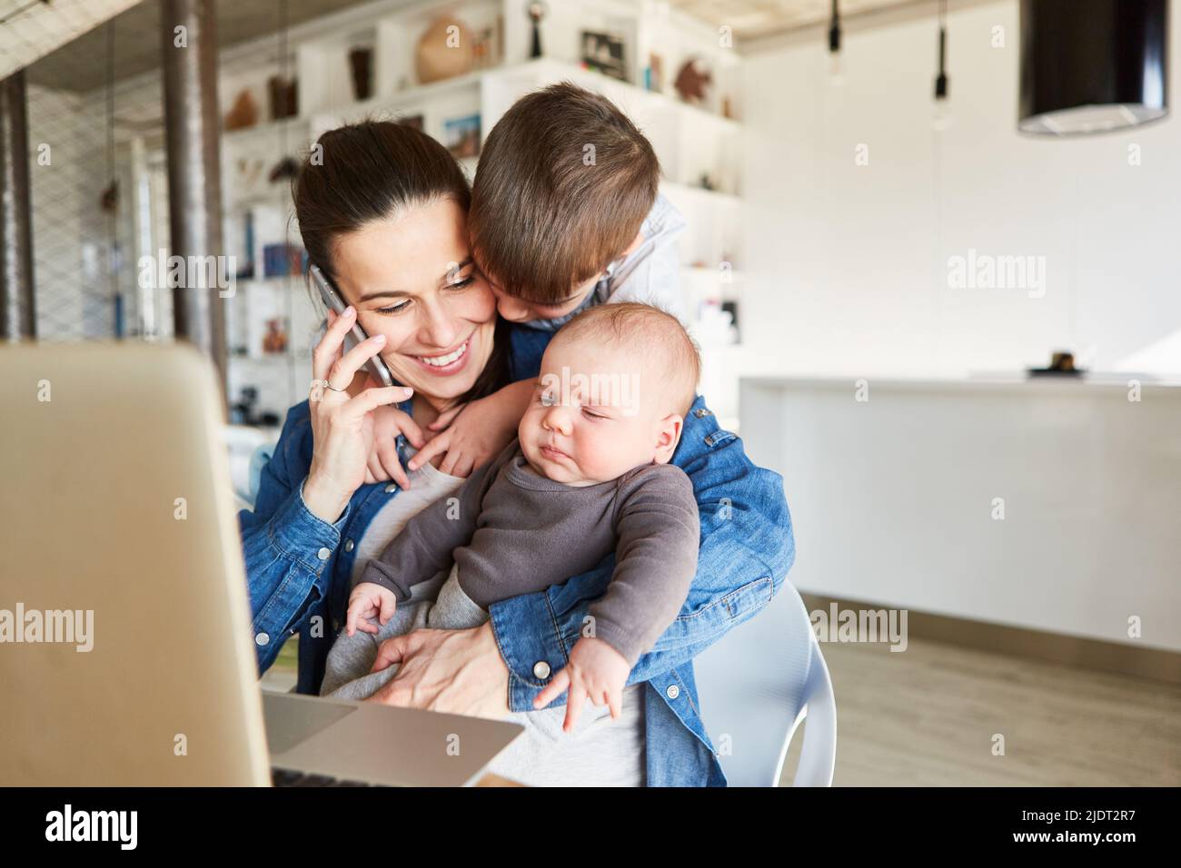 Alleinerziehende Mutter am Telefon im Heimbüro mit Baby und großem Bruder Stockfoto