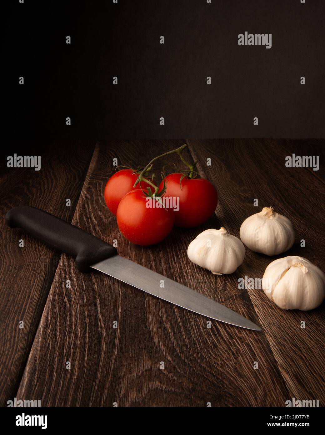 Knoblauch, Tomaten und ein Messer auf einem dunklen Holztisch mit dunklem Hintergrund und Platz für Kopien Stockfoto