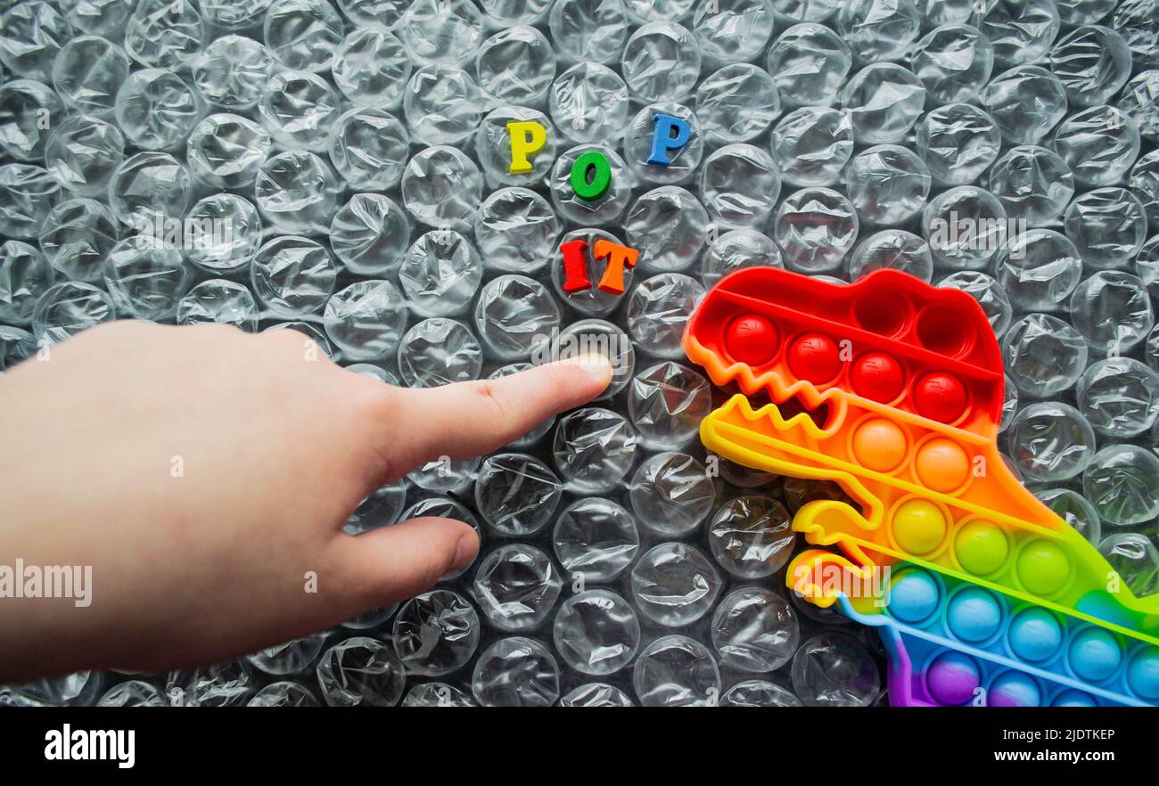 Spielzeug Pop it Dinosaurier Regenbogen Farben auf einem Luftpolsterfolie Hintergrund mit bunten Buchstaben - Pop it. Kid Finger schiebt Blase, Dino sieht es an. Kopie sp Stockfoto