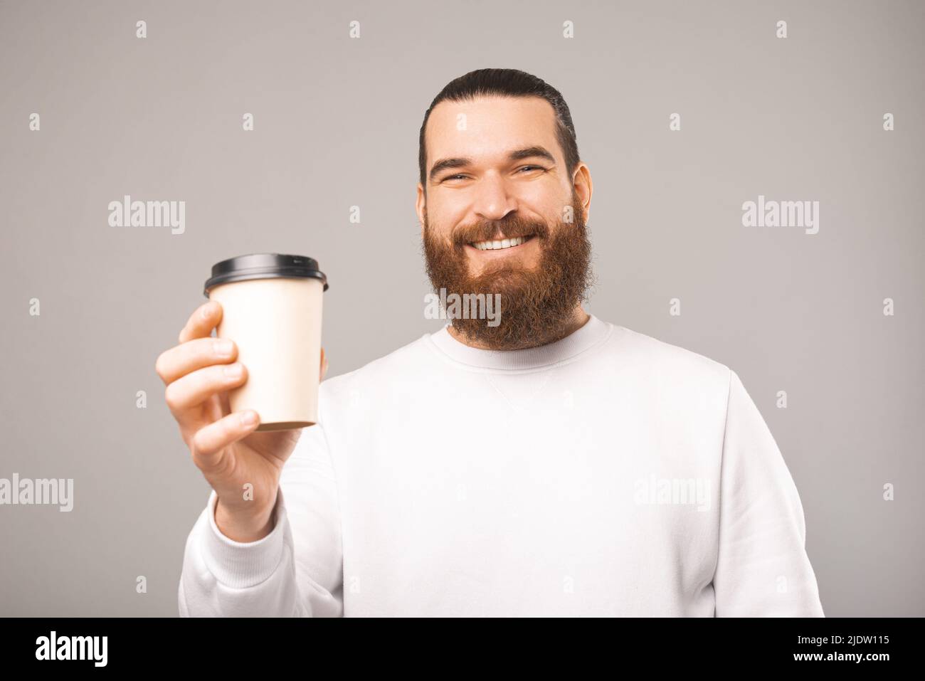 Porträt eines lächelnden bärtigen Mannes, der eine Tasse Kaffee hält und Ihnen gibt. Sudio-Aufnahme auf grauem Hintergrund. Stockfoto