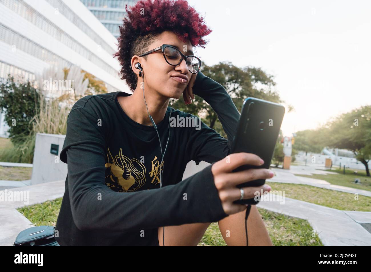Schöne junge lateinamerikanische kolumbianerin, mit rotem Afro und Brille, sitzt ruhig im Park mit Kopfhörern und Telefon bei einem Videoanruf und spricht mit ihr Stockfoto