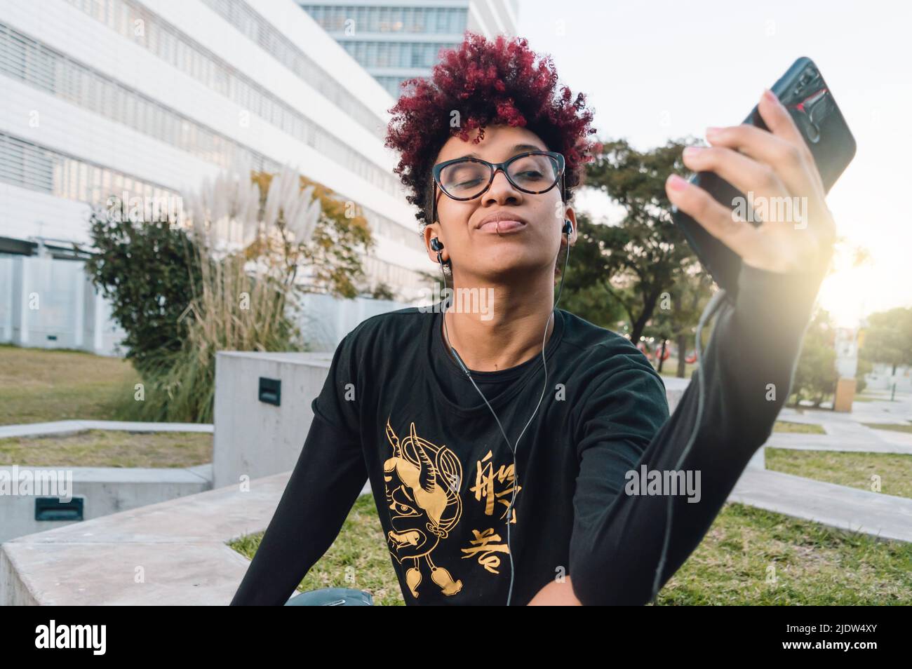 Schöne brünette kolumbianische latina Frau mit rotem Afro, Brille und gute Haltung sitzen im Park bei Sonnenuntergang, ein Selfie mit ihrem Telefon und wi Stockfoto