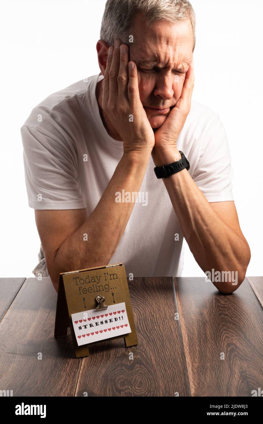 Ein älterer Mann in einem weißen T-Shirt, der den Kopf hält, auf einem dunklen Holztisch mit reinem weißen Hintergrund, der schmerzt, gestresst und besorgt aussieht Stockfoto