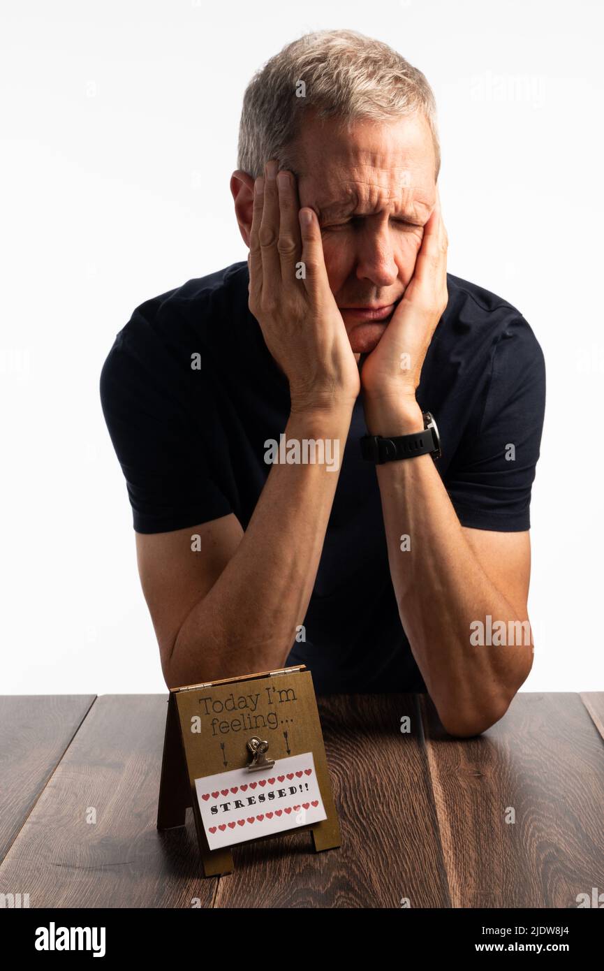 Älterer Mann in einem blauen T-Shirt, der seinen Kopf hält, auf einem dunklen Holztisch mit einem reinen, brillanten weißen Hintergrund, der schmerzt, gestresst und besorgt aussieht Stockfoto