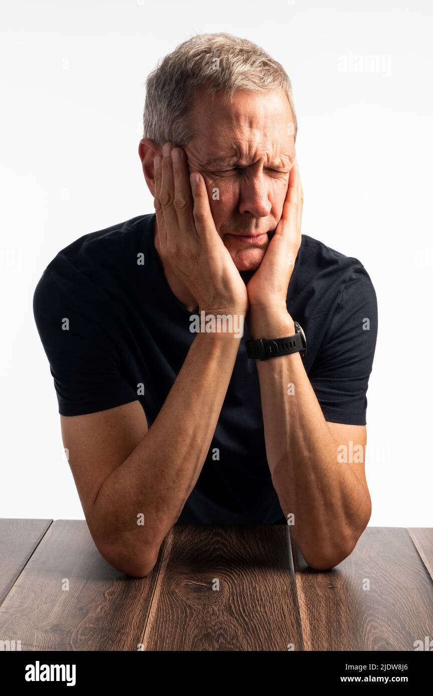 Älterer Mann in einem blauen T-Shirt, der seinen Kopf hält, auf einem dunklen Holztisch mit einem reinen, brillanten weißen Hintergrund, der schmerzt, gestresst und besorgt aussieht Stockfoto